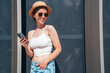 Young beautiful smiling hipster woman in trendy summer casual clothes. Carefree woman posing in the street. Positive model holds mobile phone, looks at cellphone screen, uses smartphone apps