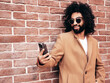 Handsome smiling hipster model.  Arabian man dressed in brown coat clothes. Fashion male posing in the street at sunny day. Holding smartphone, looks at cellphone screen, use phone apps. Scroll
