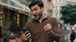Happy hopeful lucky Indian Arabian ethnic man guy businessman male young student hold mobile phone smartphone wishing hope praying crossed fingers winner yes scream victory gesture outside city street