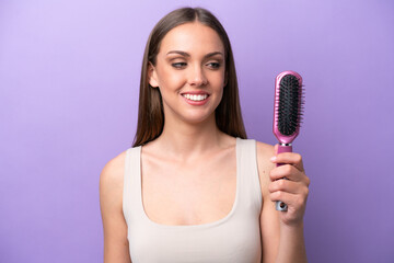 Wall Mural - Young caucasian woman holding hairbrush isolated on purple background with happy expression