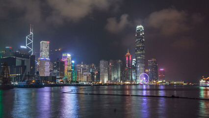 Wall Mural - Hong Kong city skyline at night over Victoria Harbor with cloudy sky and urban skyscrapers timelapse hyperlapse.