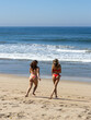 Two friends in swimwear jog playfully along a sunlit beach.