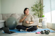 © wattana - Positive young overweight woman drinking water resting after exercising on a yoga mat at home.