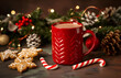 A red mug with hot chocolate inside and big white marshmallows on top, surrounded in the style of gingerbread cookies and candy canes on the table