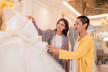 Asian bride is smiling while choosing wedding dress in modern wedding salon, Attractive designer girl using tape meter fitting on wedding dress at wedding studio.