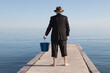 an anonymous businessman in a black suit and barefoot on a pier surrounded by water with a bucket