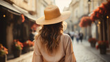 Fototapeta Uliczki - Back view of slender young woman wearing hat and coat walking down street on sunny spring day, heading to city beach to enjoy beautiful sunset. People, lifestyle, travel and vacations concept