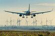A passenger aircraft taking off with wind turbines in the background, showcasing the juxtaposition of traditional and renewable energy sources.

