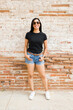 Confident latina woman standing against a brick wall, modeling a plain black t-shirt perfect for mockup designs