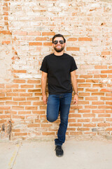 Wall Mural - Smiling hispanic man in black t-shirt and jeans posing against a rustic brick wall for a mock-up photo shoot