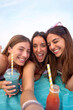 © CarlosBarquero - Vertical. Three young Caucasian women smiling taking a selfie photo looking at camera and holding fruit juices lying on a beach day. Attractive female people friends enjoying summer holidays