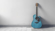Blue acoustic guitar leaning against a white wall on a wooden floor with shadows.