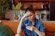 © SHOTPRIME STUDIO - Stressed woman sitting on couch looking at phone with head in hands in living room