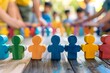 © JD - Colorful wooden figures standing on a table in focus with children and adults interacting in the background, representing teamwork and diversity