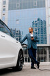 © wedmoments.stock - A business man uses a mobile phone while charging the battery of his electric car. A confident man in a business suit stands near a luxury car with a modern smartphone in his hands.