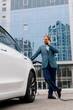 © wedmoments.stock - A young successful businessman in a stylish suit uses a telephone against the backdrop of a modern business center and a modern electric machine. Concept of electric vehicles, business and style.