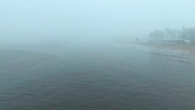 The Camera Pans Backward, Revealing A Misty Beach Veiled In Dense Fog. As It Retreats, A Small Pier Comes Into View, Where A Man Casts His Fishing Line Amidst A Group Of Seagulls Fluttering Overhead.