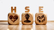 The letters word text abbreviation acronym HSE above three wooden blocks, with symbols on each block representing the different aspects of health, safety, and the environment.