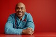 © Riccardo - Confident male doctor sitting at a table in clinic attire, against a vibrant red background, exuding cheerfulness