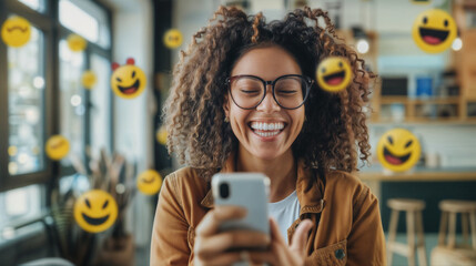 A young smiling black woman holding a mobile phone in the office with emoji smiles flying around her. The person receives a pleasant message