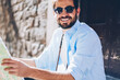 Cropped portrait of happy bearded tourist in stylish sunglasses holding travel map for searching showplaces in hands.Positive tourist in casual wear smiling at camera while sitting on street
