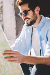 Wall Mural - Happy casual dressed male tourist with beard and stylish sunglasses looking at map to determine route of interesting showplaces sitting outdoors on street.Positive young man searching right direction