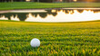 White golf ball on picturesque green golf course near pond, at sunset. Pitch is perfectly prepared, grass is neatly trimmed. Reflection of sun rays on surface of ball and texture of grass. Copy space