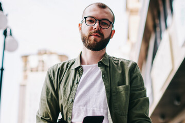 Portrait of young serious caucasian man in eyeglasses looking in camera while waiting friend on street and dialing number on cellphone, hipster guy holding smartphone in hands and typing text message