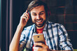 Cheerful bearded hipster guy choosing song in music app to listen in modern headphones connected via bluetooth to smartphone.Positive male blogger chatting while enjoying audio from player on cellular