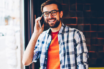 Wall Mural - Portrait of happy bearded young man dressed in trendy shirt smiling at camera while talking with friend about funny news during phone call on smartphone.Cheerful hipster guy communicating on cellular