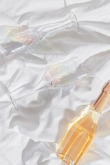 Wall Mural - Rainbow color shining champagne glasses and white sparkling wine bottle on bed, on white blanket background. Lifestyle aesthetic photo, star filter. Romance meeting, romantic holiday concept.