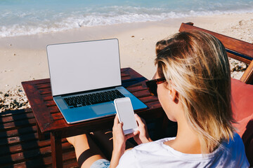 Wall Mural - Back view of blonde female blogger checking notification on smartphone working on ocean beach using technology, businesswoman spending time in journey on distance job using laptop with blank screen