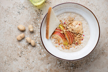 Wall Mural - Bowl of oatmeal with peanut butter, fresh apple and granola, horizontal shot on a beige granite background with space, high angle view