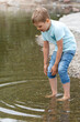 A small cautious boy in blue clothes wades into the water and tries to pull up his pants so they don't get wet