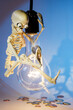 A funny, smiling skeleton swinging on a light bulb, in light blue background, kindly invite to save electricity because, he already saved a lot of coins. Free space for text. Vertical