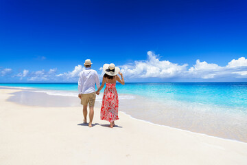 Happy holiday couple with sunhats walks down a tropical paradise beach with turquoise sea and golden sand