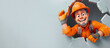 Cartoon cheerful male builder in an orange suit and helmet looks out of a hole on a gray background with copyspace