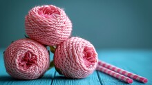   Three Pink Yarn Skeins Adjacent To A Pair Of Pink Knitting Needles On A Blue Table Against A Green Backdrop