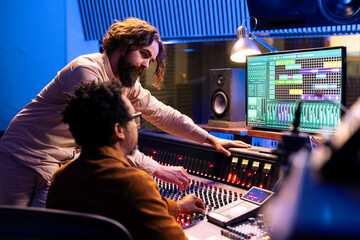 Fototapete - Skilled artist working with audio technician to edit recorded songs in control room, pressing sliders and levers on panel board. Team of singer and sound designer creating records in studio.