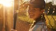 A girl in a baseball uniform holding a baseball bat stands smiling and looks at the camera with sunlight shining behind her