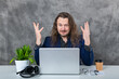 Stylish man with long hair in blue suit striking poses by laptop and accessories