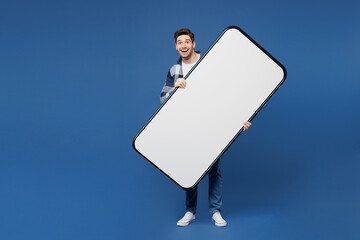 Wall Mural - Full body young man he wears shirt white t-shirt casual clothes hold in hand big huge blank screen area mobile cell phone smartphone look camera isolated on plain blue background. Lifestyle concept.