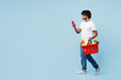 © ViDi Studio - Full body young Indian man wear white t-shirt casual clothes hold basket for takeaway with food products, mobile cell phone isolated on plain blue background. Delivery service from shop or restaurant.