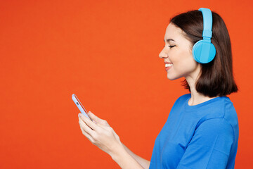 Canvas Print - Side view young smiling fun cool happy woman she wear blue t-shirt casual clothes listen to music in headphones use mobile cell phone isolated on plain red orange background studio. Lifestyle concept.