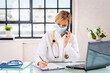 Female doctor with surgical mask sitting in doctor's room and using smartphone