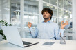 © Liubomir - A young man with curly hair meditates at his office desk, eyes closed and hands in a mudra pose, promoting mental health and stress management in a professional environment.
