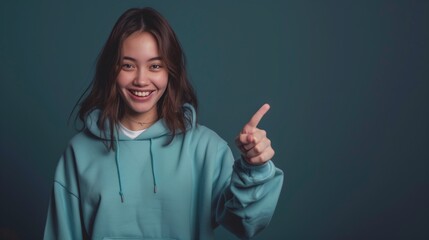 Sticker - Young Asian woman in a teal hoodie smiling and pointing with one finger, dark background.