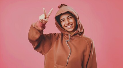 Wall Mural - Joyful young woman in a brown hoodie making peace sign against a pink background.