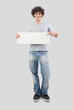 Young man, smiling and handsome, pointing with finger to show a blank white signboard, isolated on gray background. Placard copy space for text or logo, banner for advertising or promotional messages