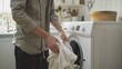 Man in gray shirt folding white laundry in front of white washing machine in kitchen.
